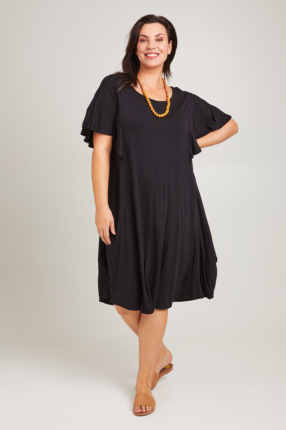 Model Poppy wearing size 16 of the Tahiti Frill Dress in colour Black (Black), from the front, showing the whole outfit